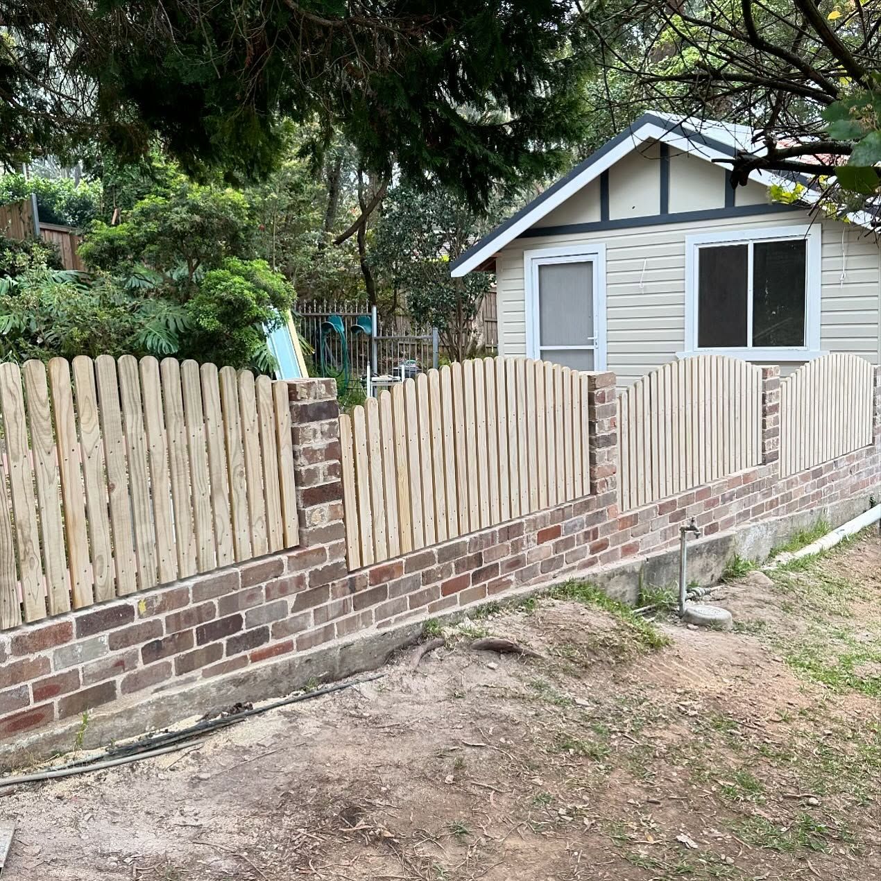 A wooden picket fence with a scalloped top sits atop a red brick base in front of a light-colored shed.