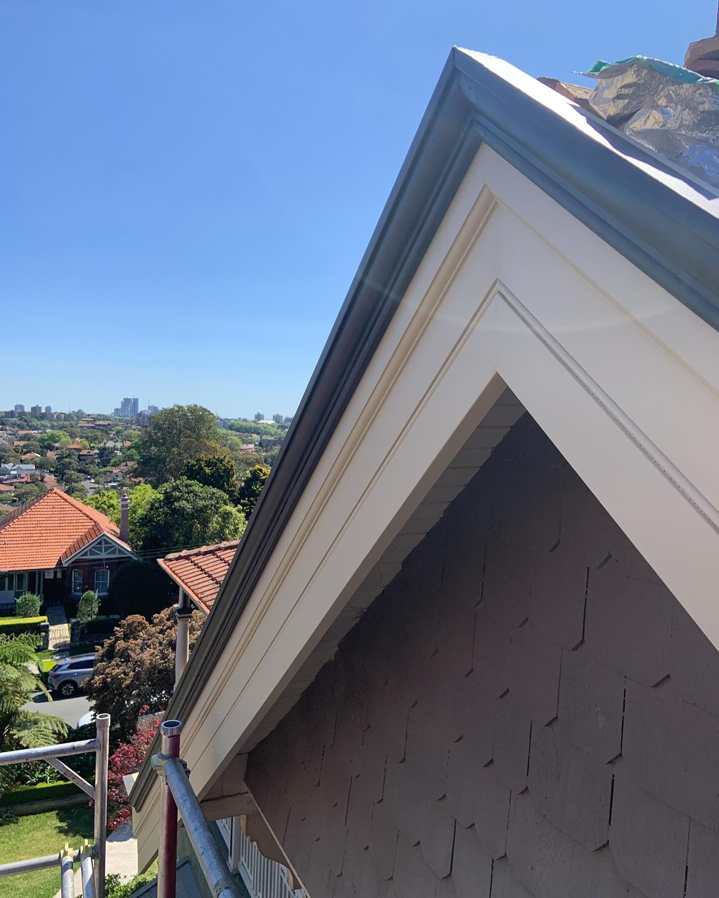 A high-angle view of a brown gable wall with peeling paint, adjacent to white roof trim, overlooking a neighborhood.