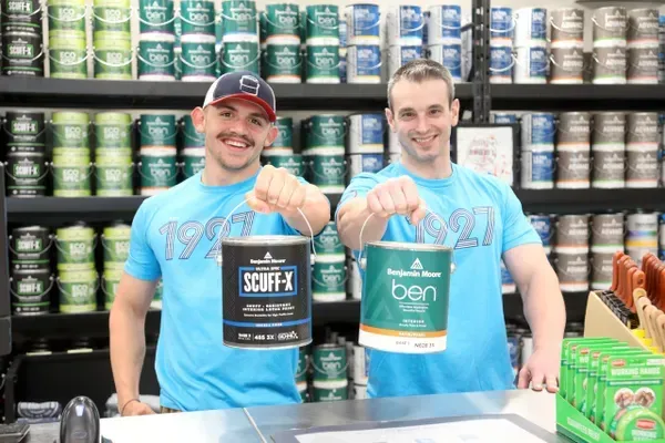 Two men are standing next to each other in a paint store holding buckets of paint.