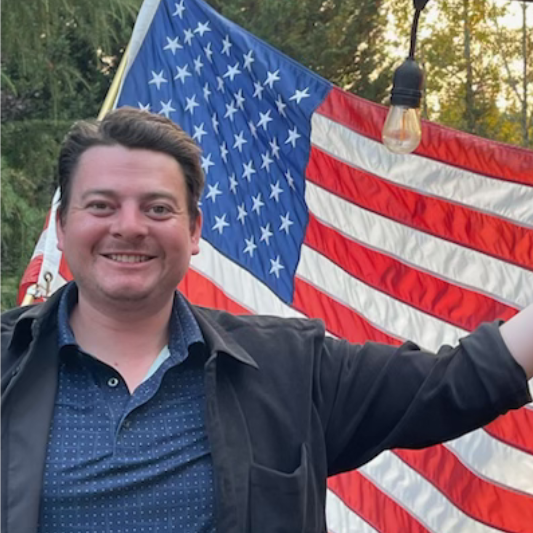 Smiling man in a dark jacket posing in front of a large American flag outdoors