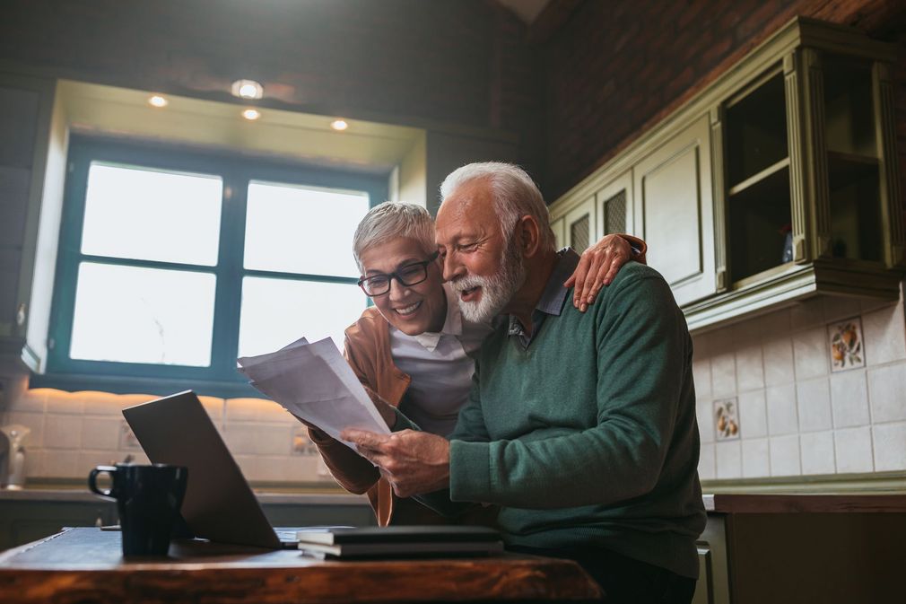 An elderly couple is sitting at a table looking at papers and a laptop.