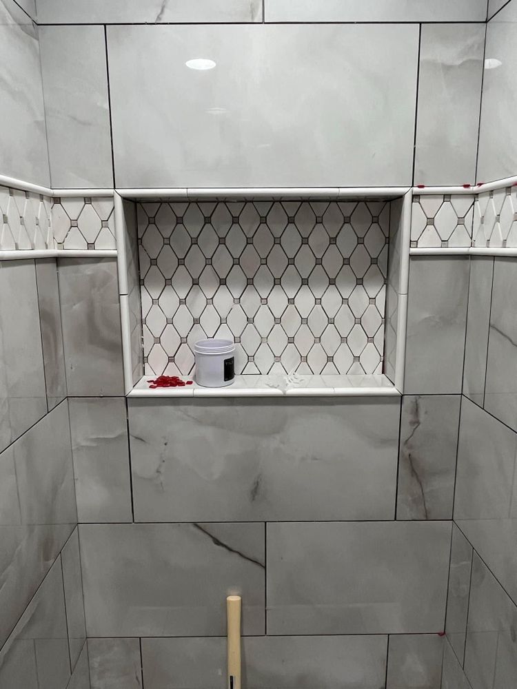 Gray tiled shower with built-in niche holding a soap dispenser and red item, and a wooden-handled brush below
