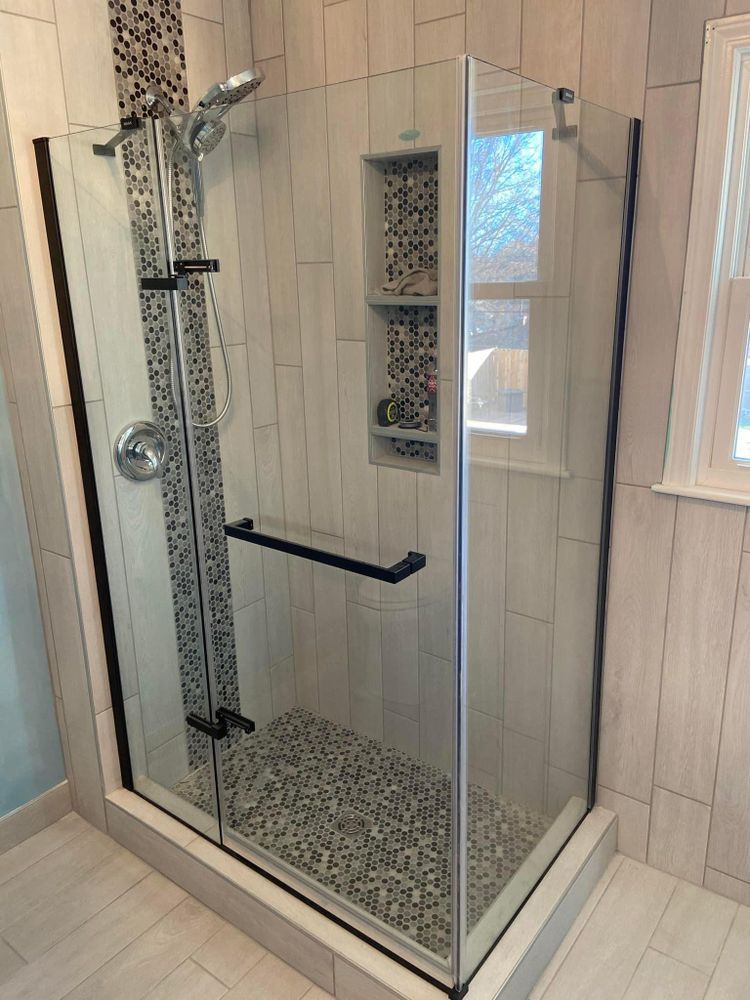 Glass corner shower with pebble tile floor and chrome fixtures in a light wood-paneled bathroom