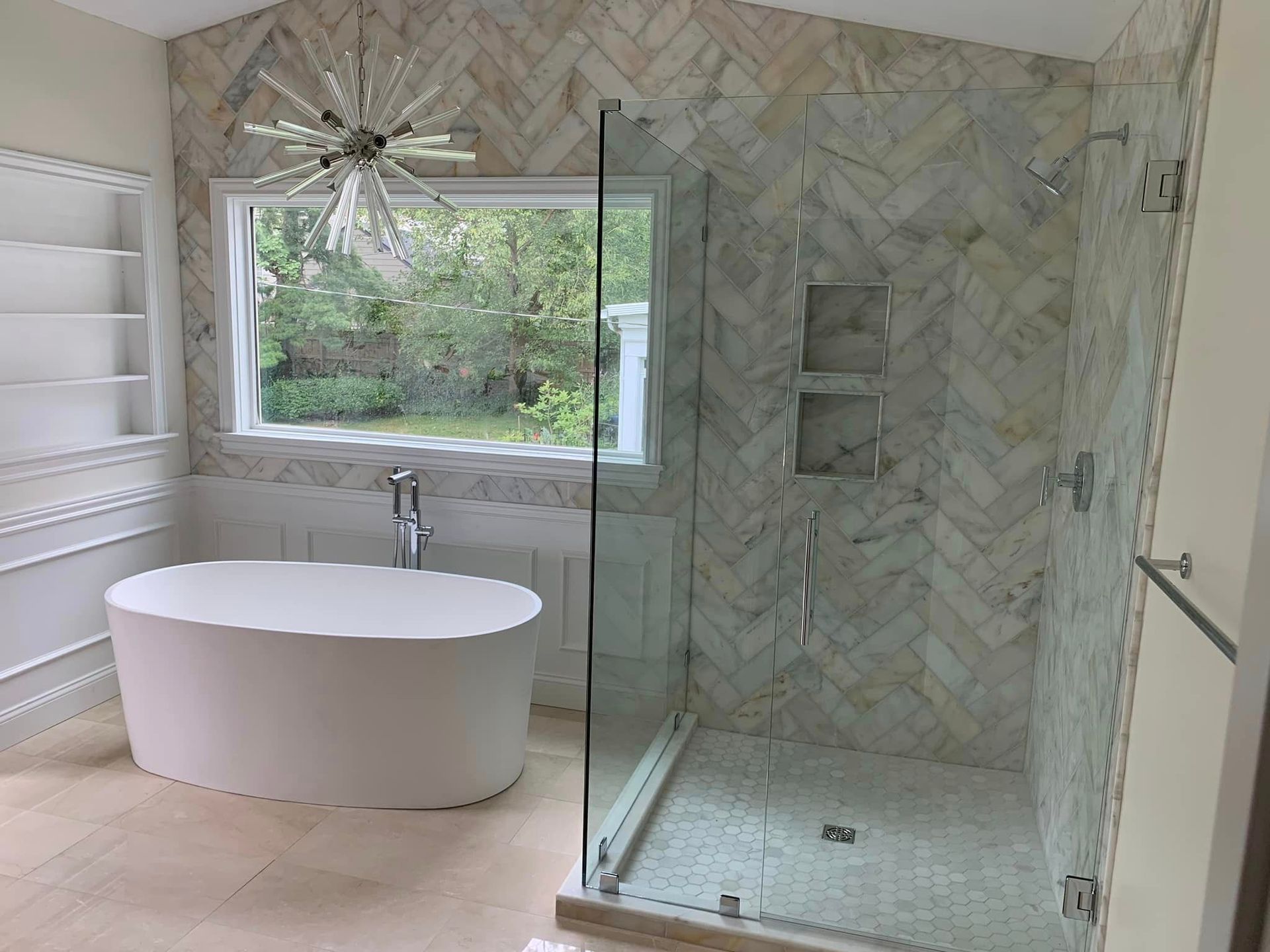 Modern bathroom with freestanding tub, glass shower, marble walls, and large window overlooking greenery