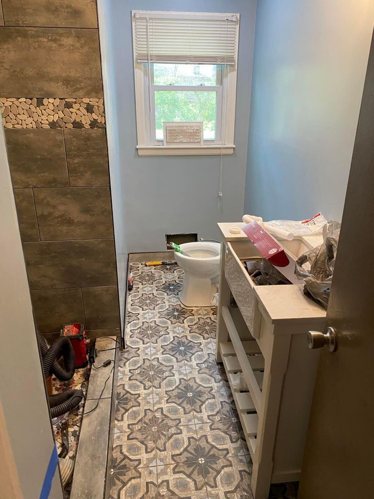 Narrow bathroom under renovation with patterned floor tile, partially tiled shower, vanity, bucket, and window.