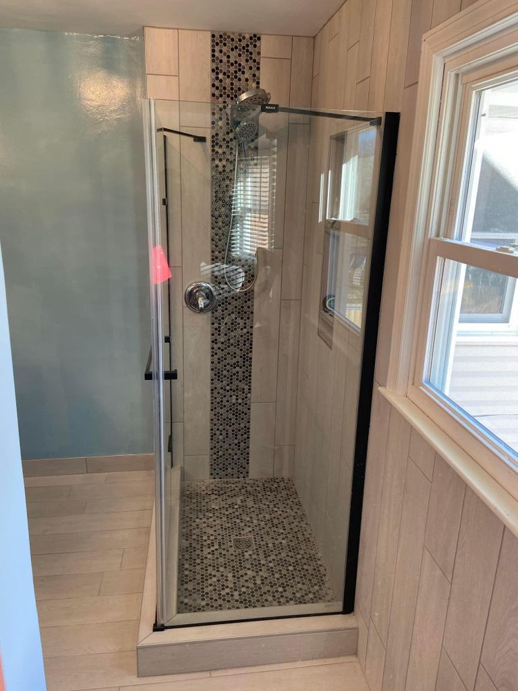 Bathroom with glass shower enclosure, pebble floor, and light wood paneling beside a window