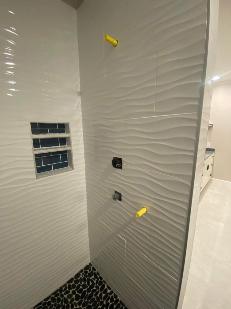 White tiled shower stall with two wall fixtures, a recessed niche, and black pebble floor tiles.
