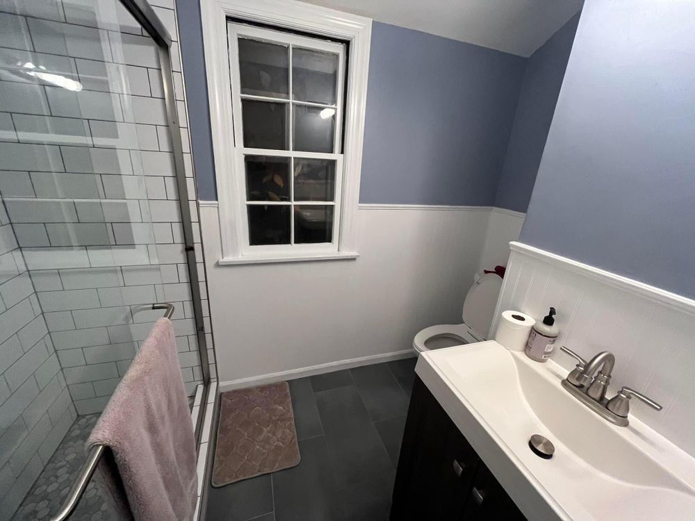 Small bathroom with white tile shower, window, dark floor, and vanity sink.