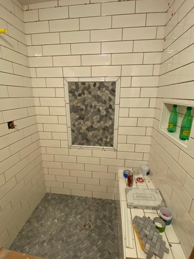 Small tiled shower with gray mosaic floor, frosted window, and bathroom items on a shelf.