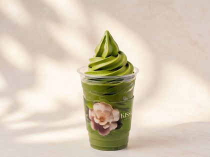 Clear cup of green soft-serve topped with swirls, decorated with a flower sticker, on a light background