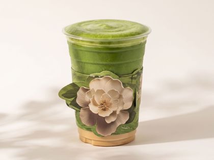Green smoothie in a clear cup with a beige flower decoration and lid on a light background