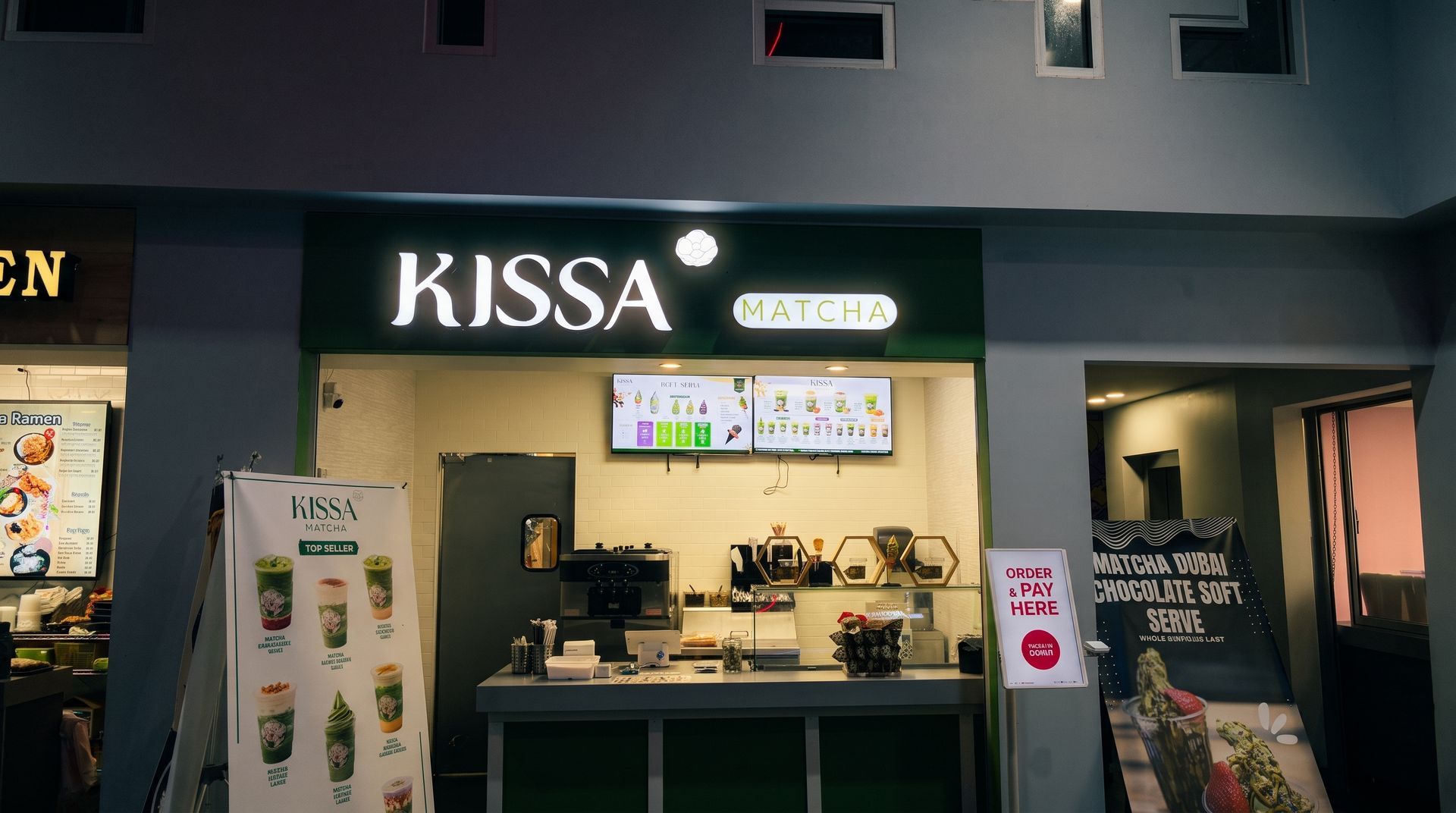 KISSA café storefront at night, with lit menu screens and a service counter inside