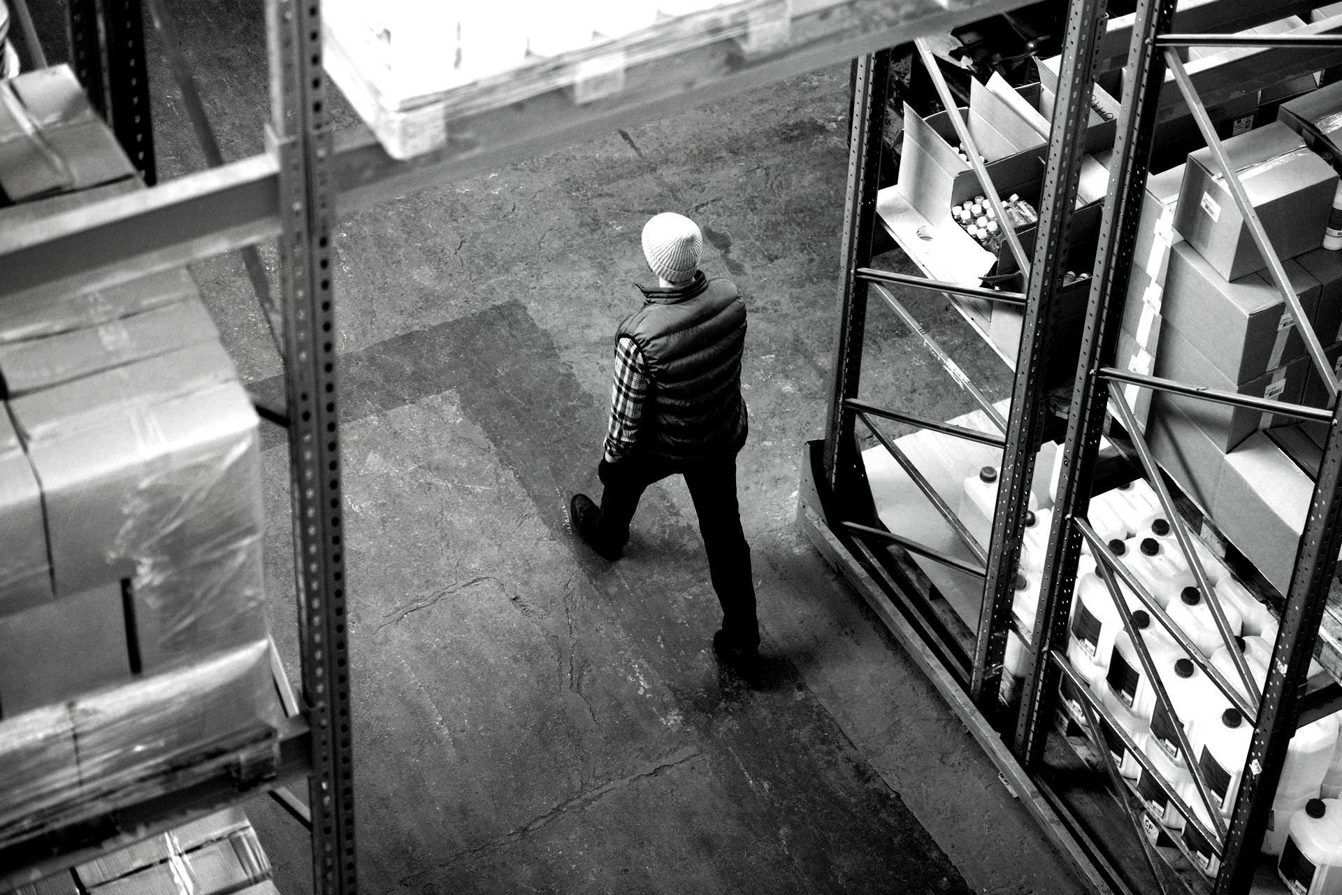 Person walking through a warehouse aisle between stacked pallets and metal shelving.