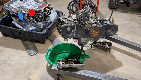 Disassembled car engine parts on a shop floor, with a green transmission housing in the foreground.