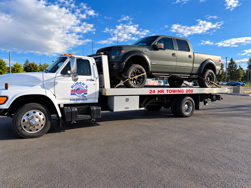Tow truck carrying a black pickup truck on a flatbed in a parking lot under a blue sky