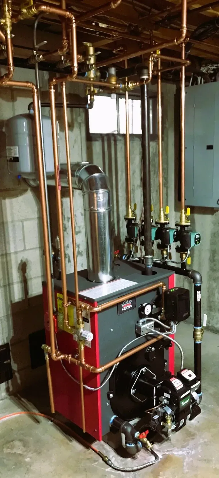 Basement boiler room with red-and-black heating unit, pipes, valves, and exposed ceiling ducts