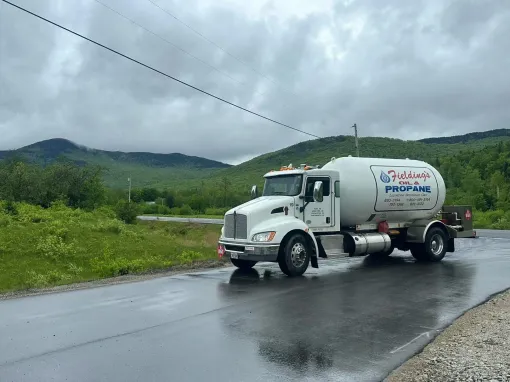White tanker truck on a wet rural road beside green hills under cloudy skies.