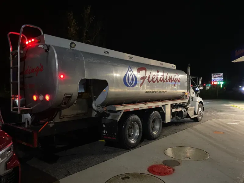 Silver fuel tanker truck parked at night on a road, with red taillights glowing and road signs in the distance.
