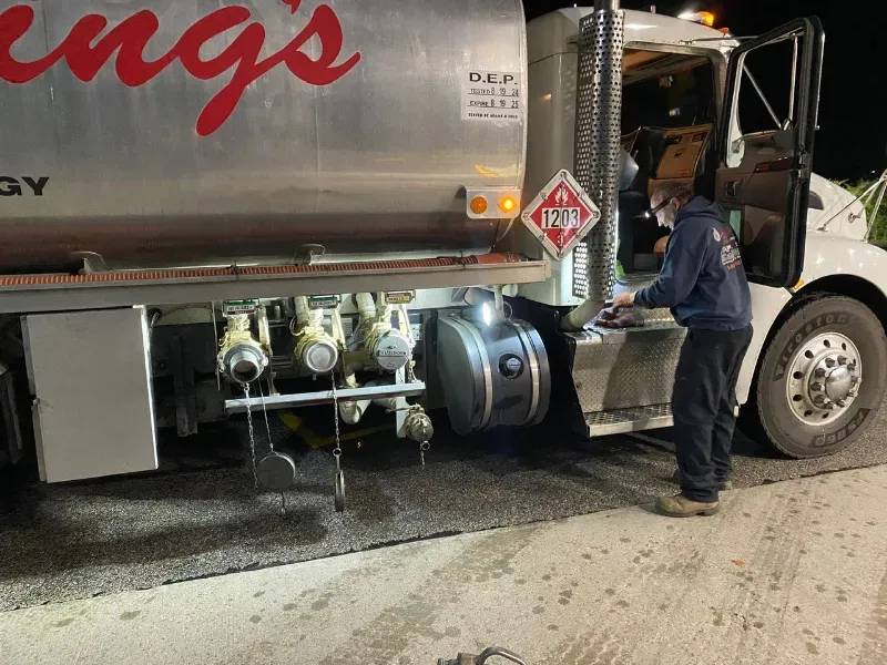 Worker inspecting the underside of a tanker truck at night on a wet roadside.