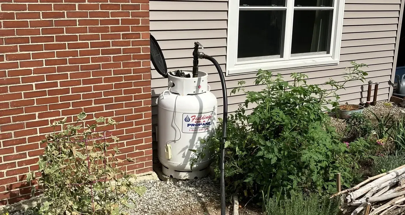 White propane tank beside brick and siding house, surrounded by bushes and gravel.