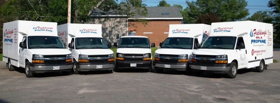 Five white delivery trucks parked in a row in front of a building, with trees in the background.