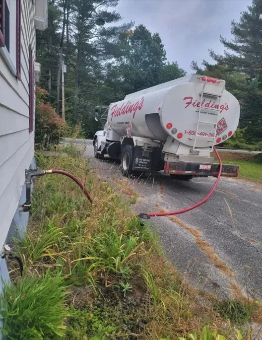 White fuel tanker truck on a wet driveway near a house, with a red hose on the ground and trees in b