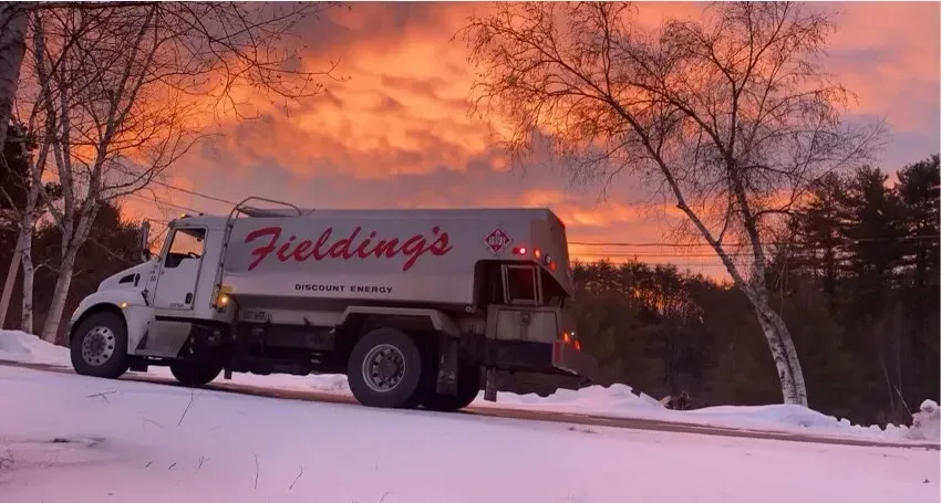 Red fire hose connected to house spigot, leading across lawn to a fuel truck near trees.