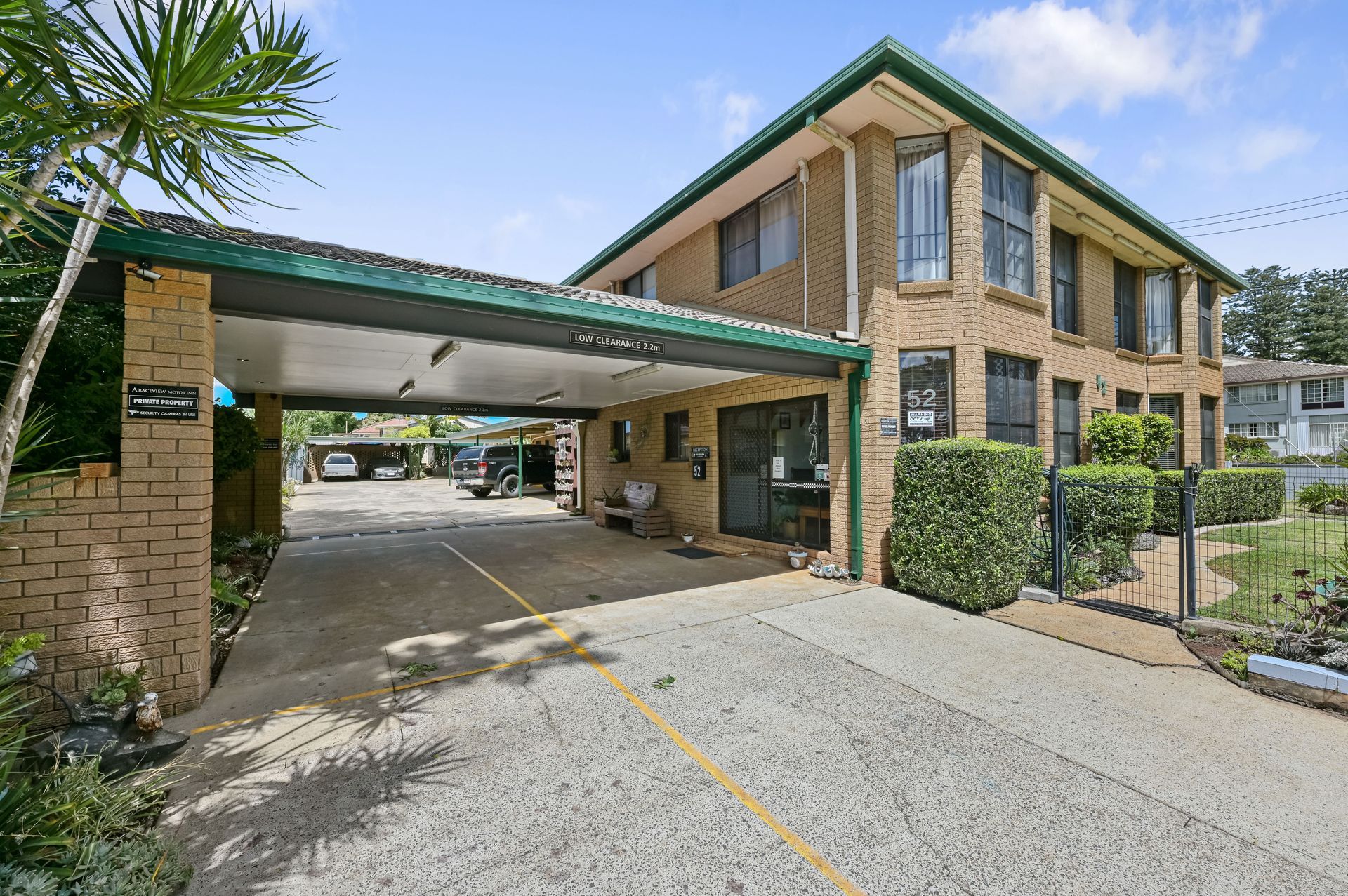 Brick apartment building with covered carport, driveway, and small fenced courtyard on a sunny day