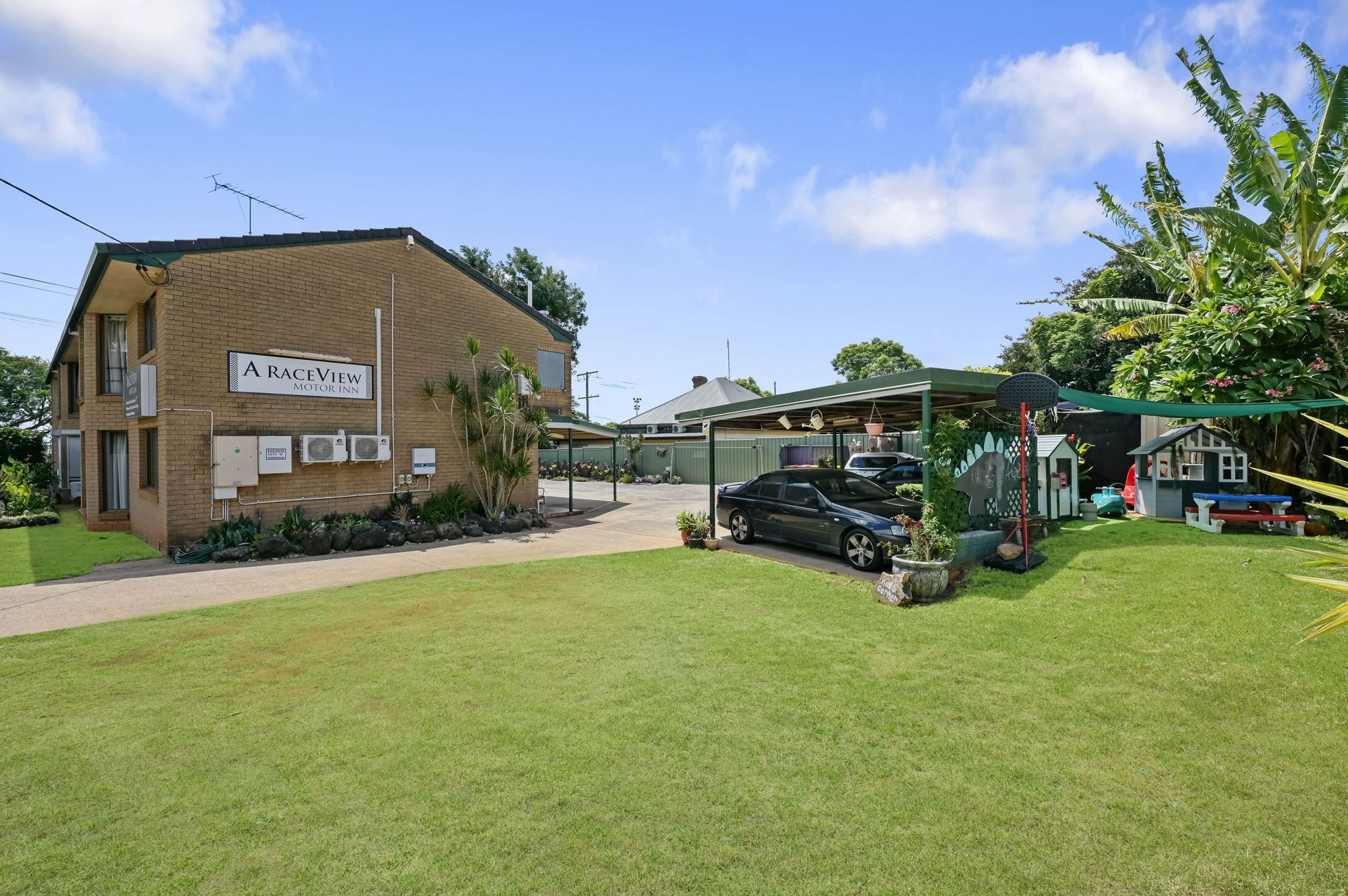Brick building with a sign, parked cars, and a large grassy yard under a blue sky