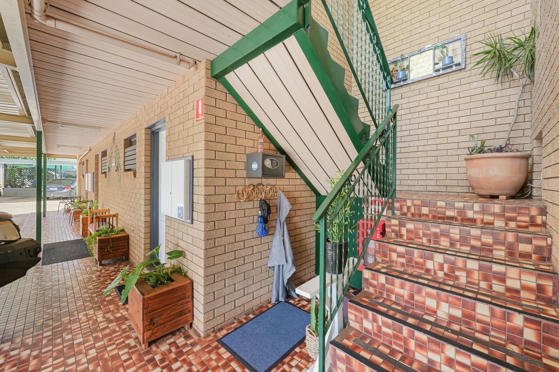 Apartment walkway with brick stairs, green railing, potted plants, and a covered entry door