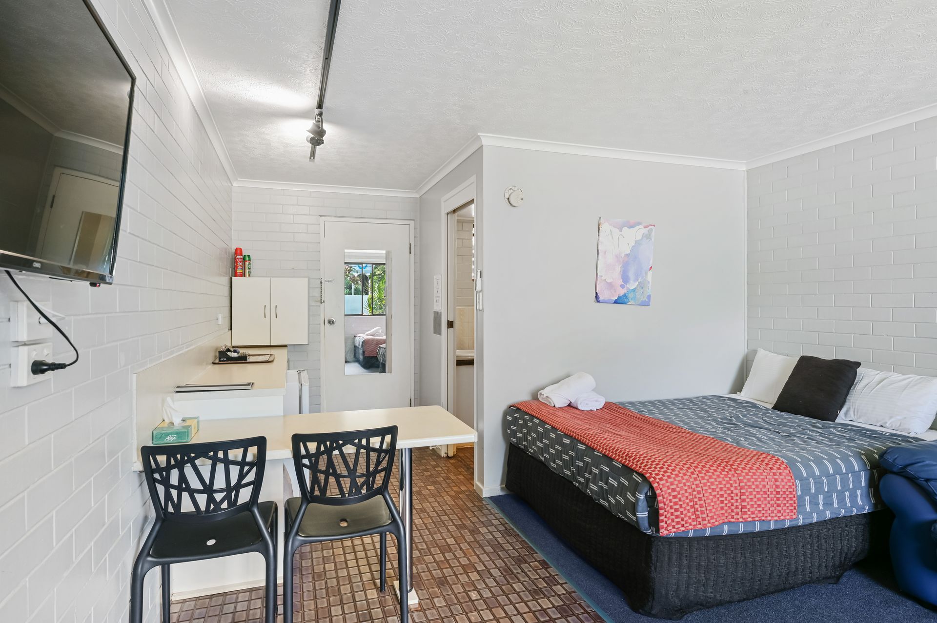 Small studio room with a bed, kitchen table, two chairs, wall TV, and kitchenette.