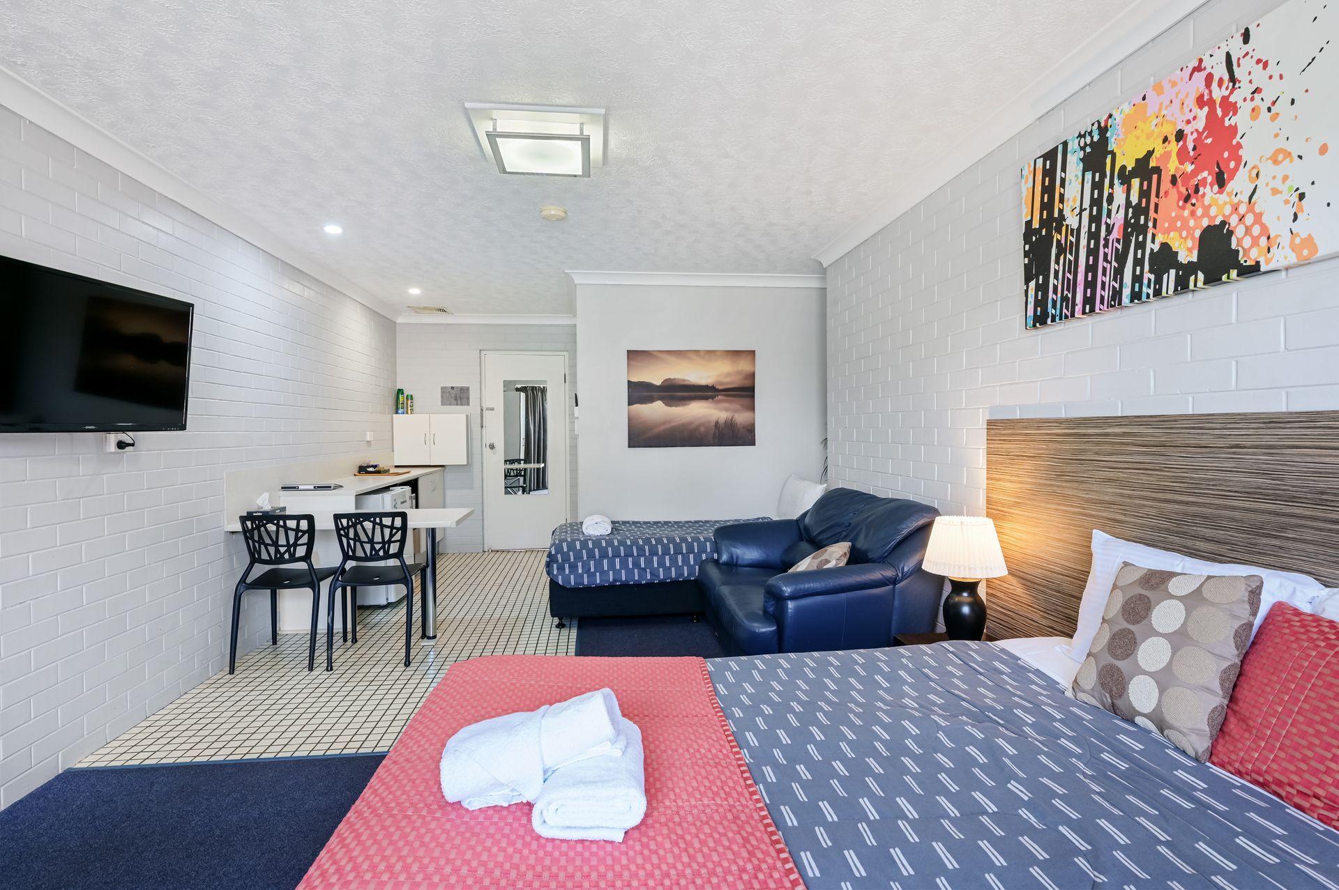 Modern hotel room with bed, blue sofa, desk, wall-mounted TV, and abstract wall art