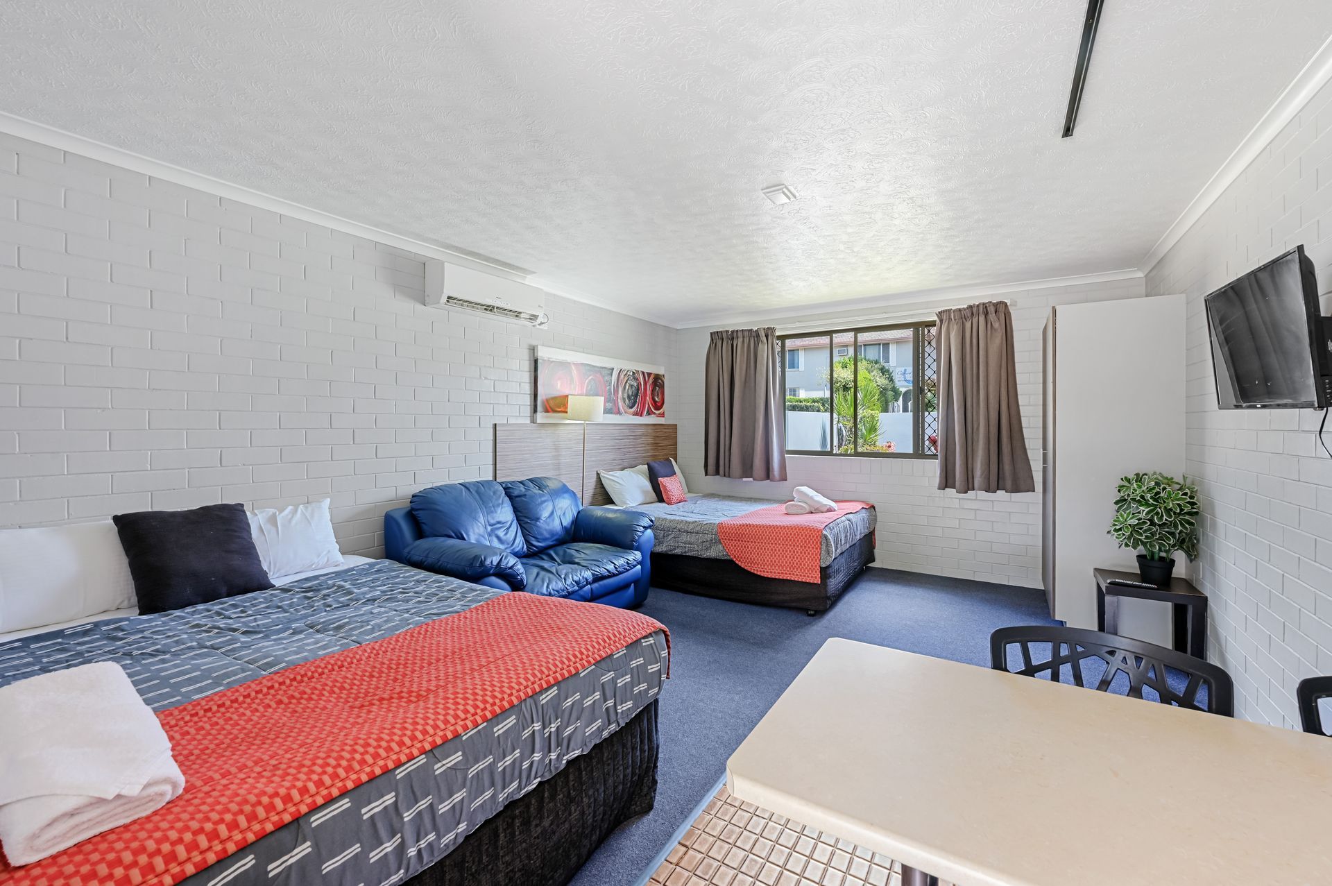 Bright bedroom with two beds, blue couch, desk, window, and wall-mounted TV