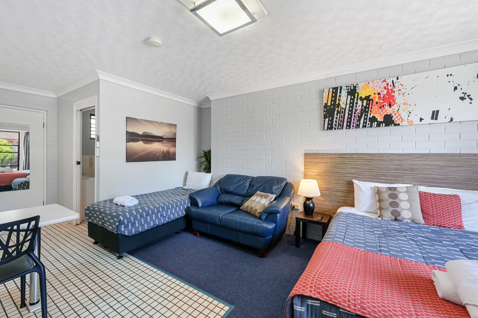 Bright bedroom with three beds, blue and red bedding, patterned rug, and wall art under a skylight.