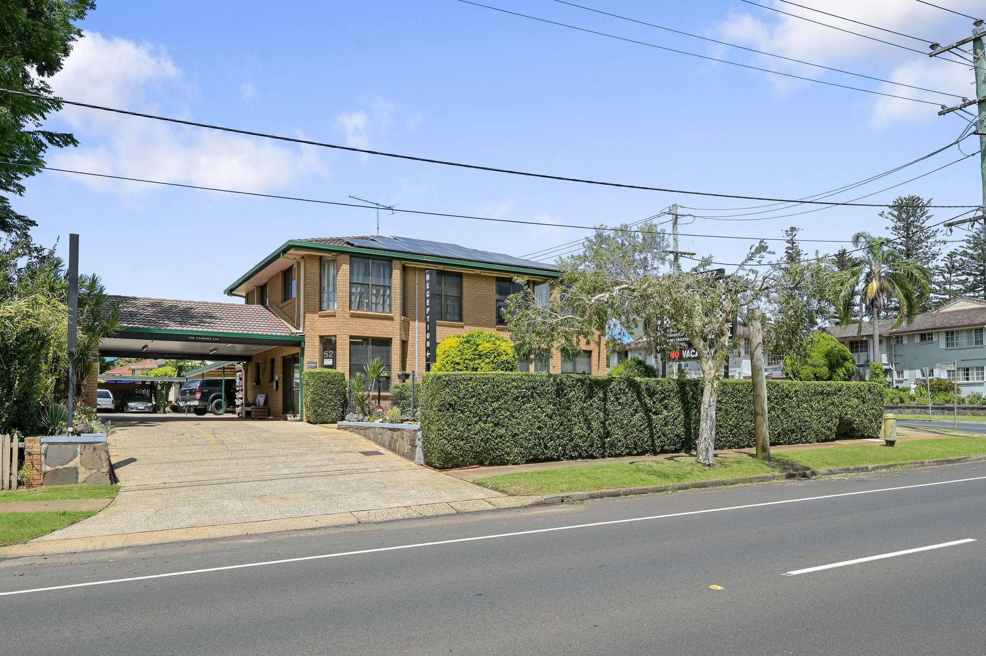 Two-story brick house with hedge-lined yard and driveway beside a road under a blue sky