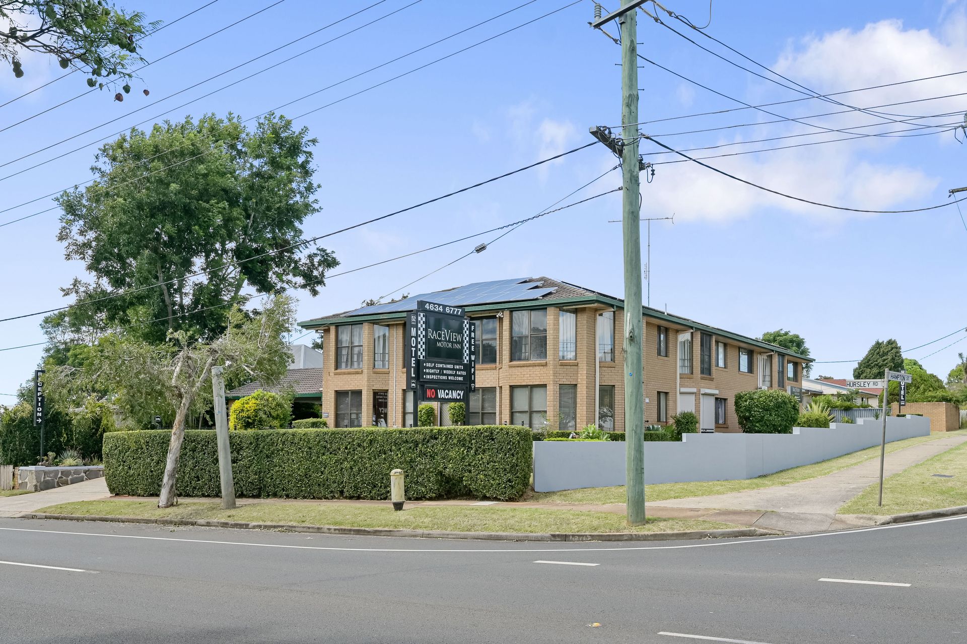 Corner view of a two-story brick building behind a hedge, with power lines and a road in front