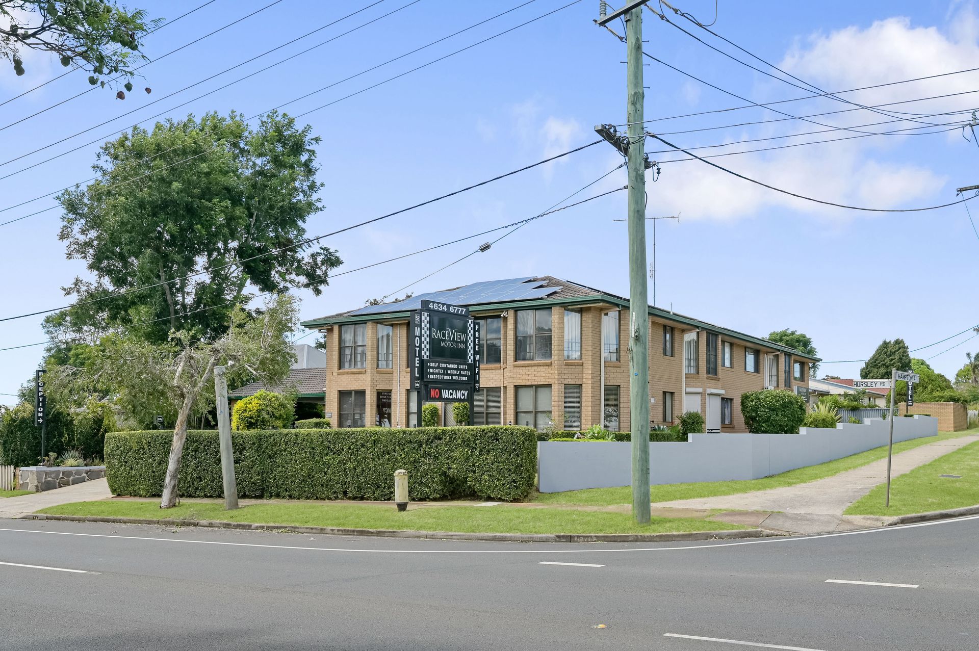 Modern apartment building on a corner lot with hedges, trees, and power lines under a blue sky