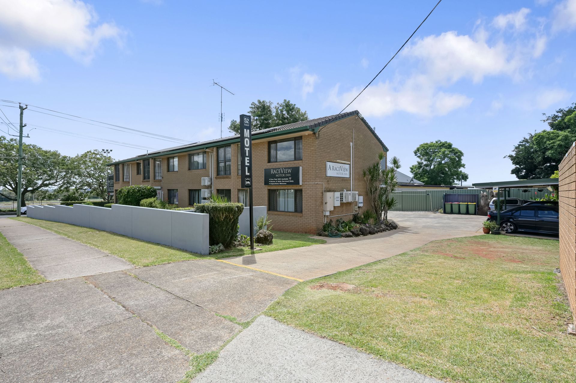 Apartment building with brown brick facade, paved driveway, lawn, and parked car under blue sky