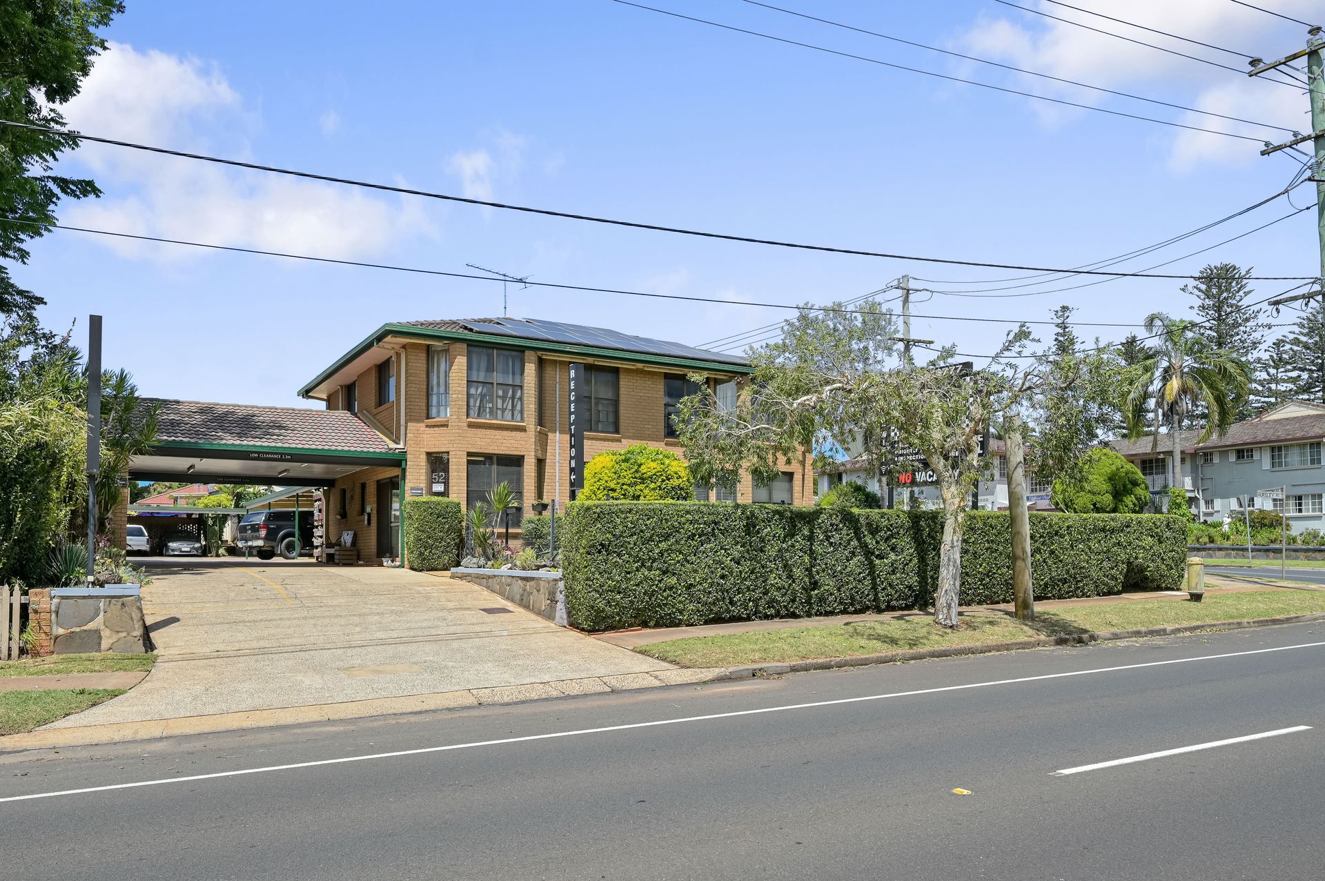Brick building with hedges and a driveway beside a road under a blue sky