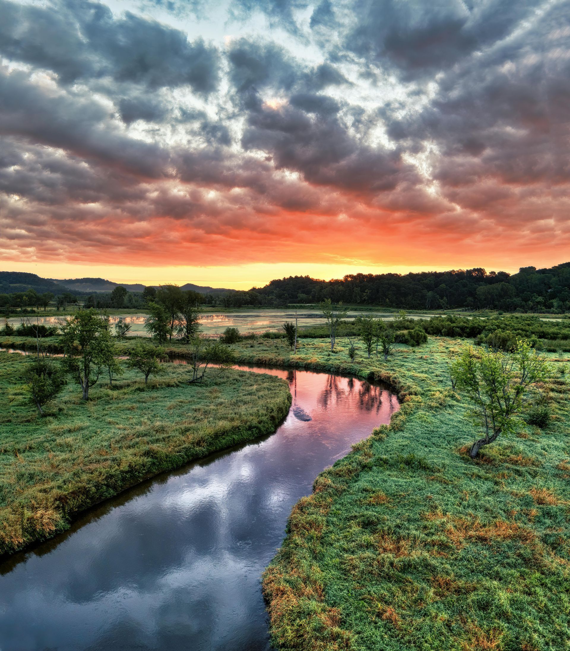 Sunset over a winding river through green fields beneath dramatic, cloud-filled sky