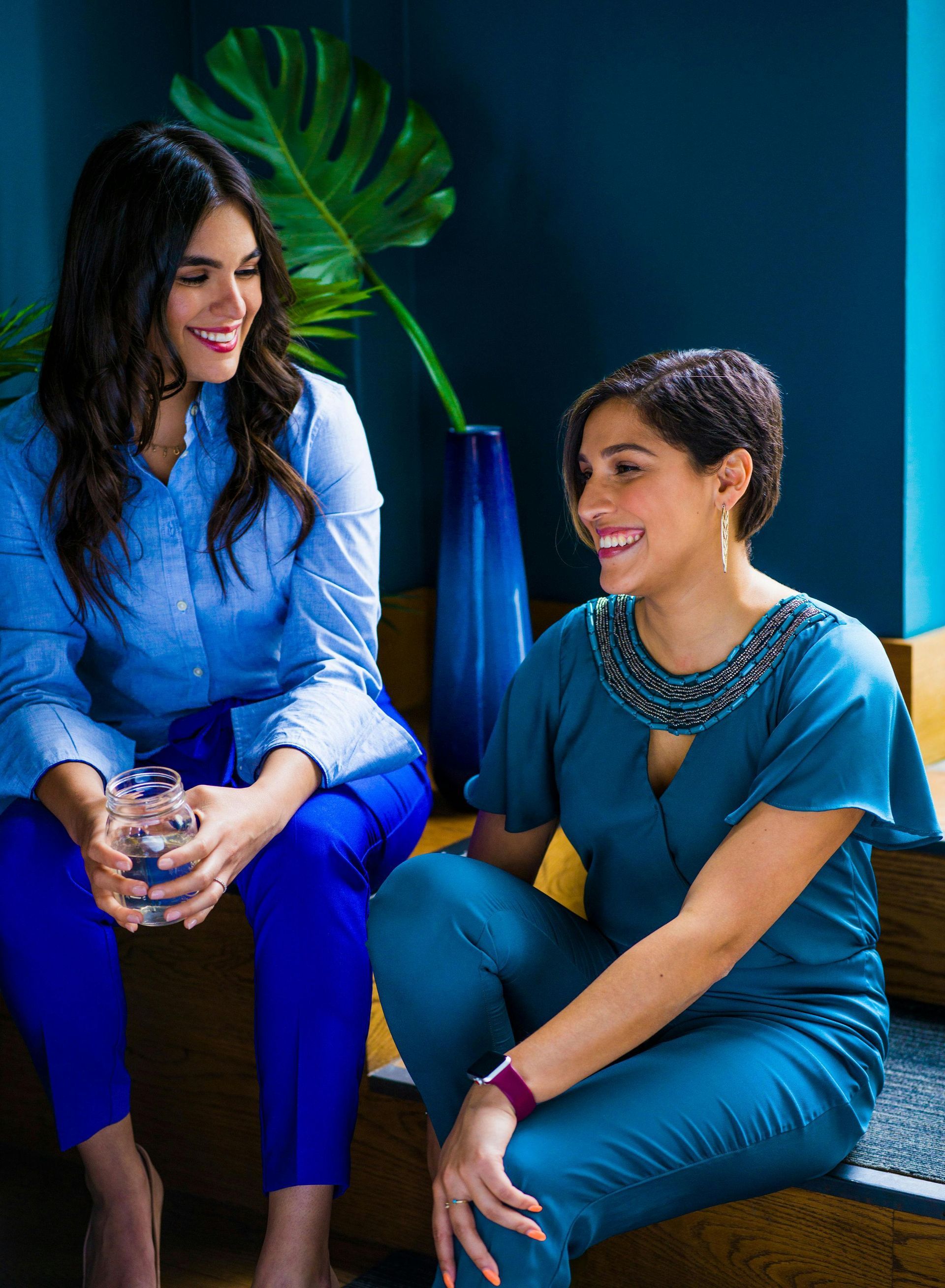 Two women in blue outfits smiling and talking while seated in a cozy indoor room
