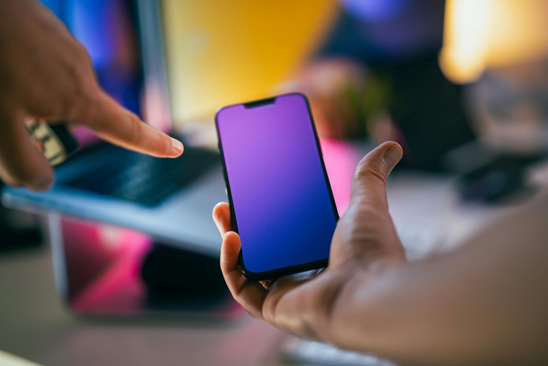 Hands holding a smartphone with a purple screen, with a finger pointing at it in a blurred indoor setting