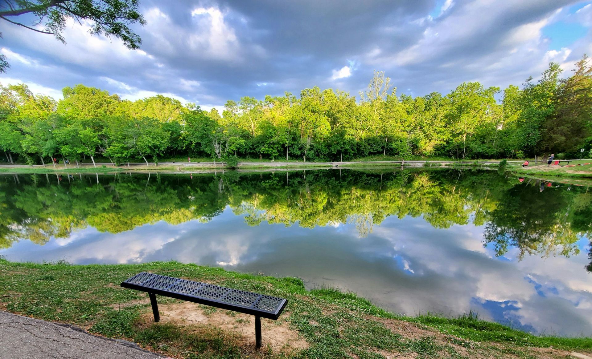Park bench beside a calm pond reflecting green trees and a cloudy sky.
