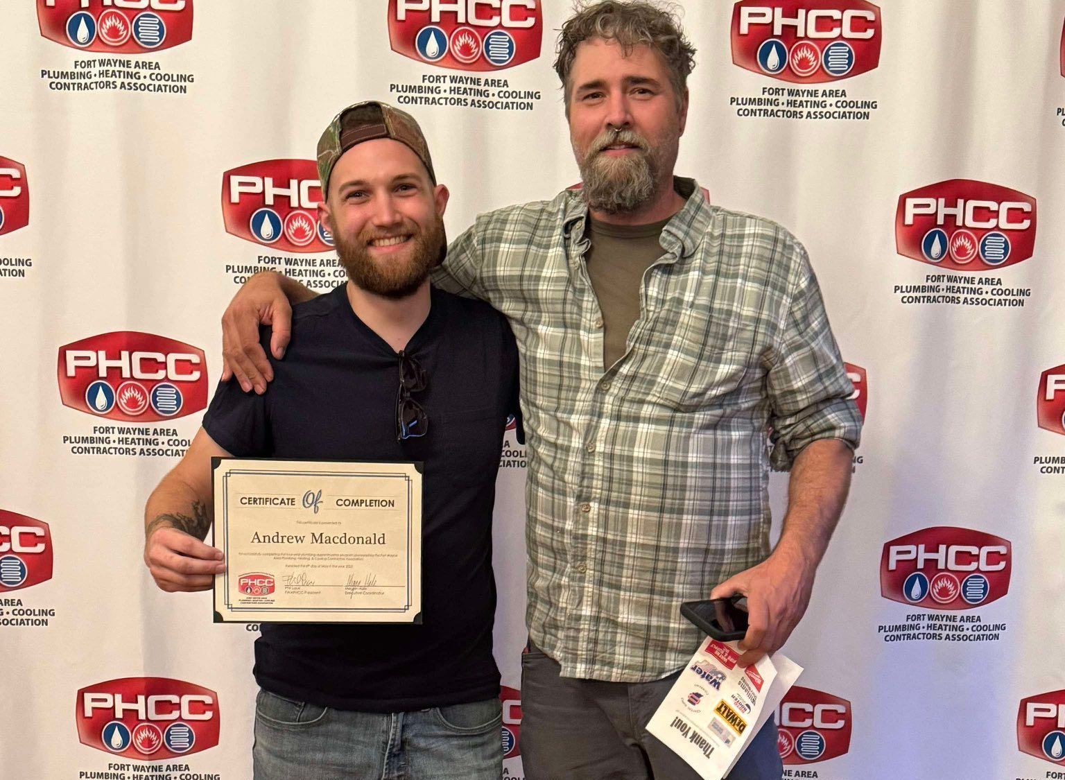 Two men pose at a PHCC event, one holding a certificate in front of a logo backdrop.
