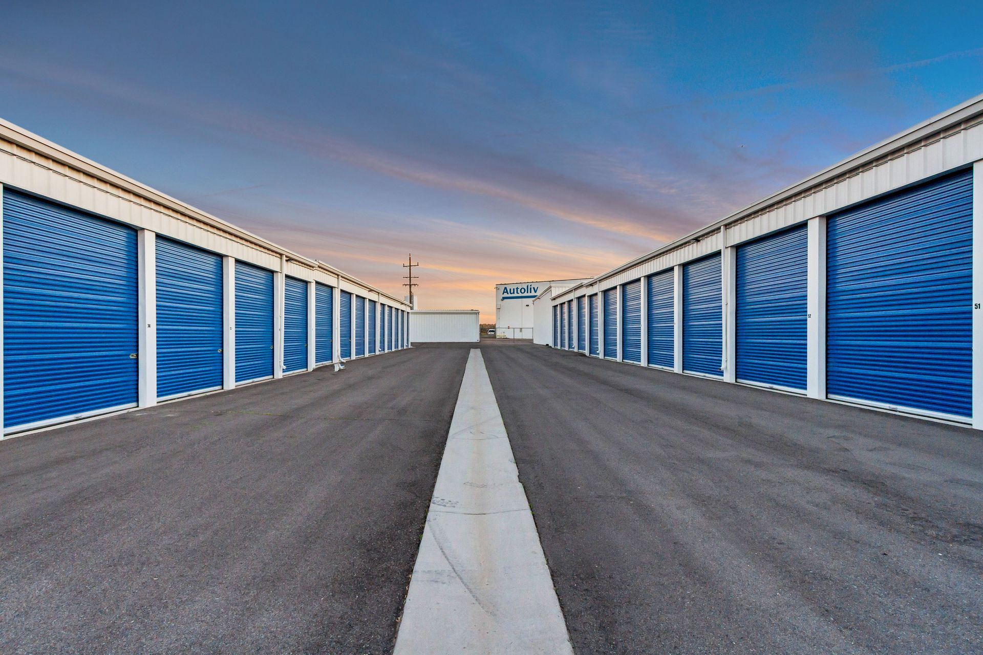 Rows of blue storage units with white trim, asphalt road, and dusk sky.