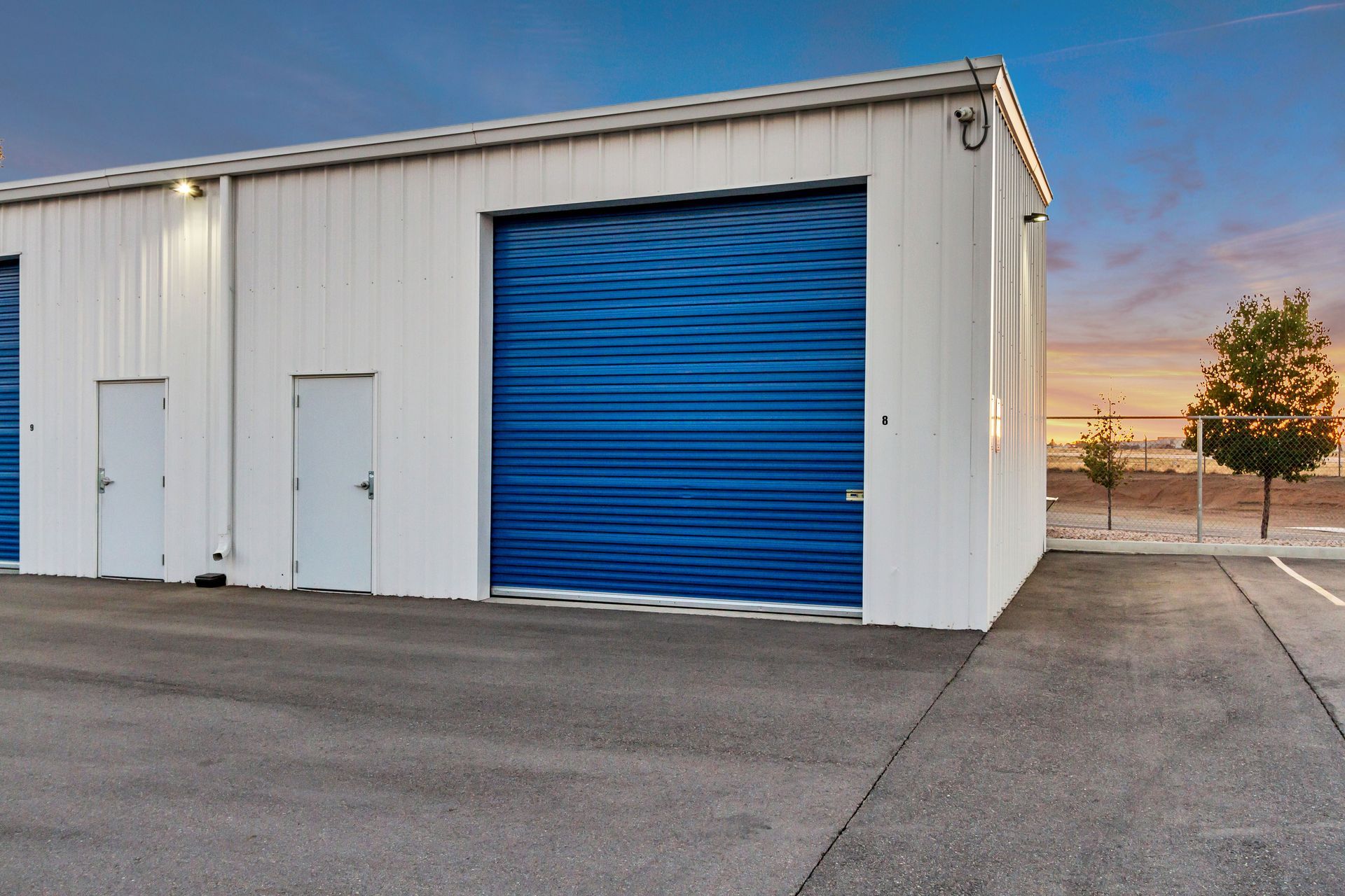 Storage units, white metal building with blue roll-up door and two white doors, asphalt parking area, sunset sky.