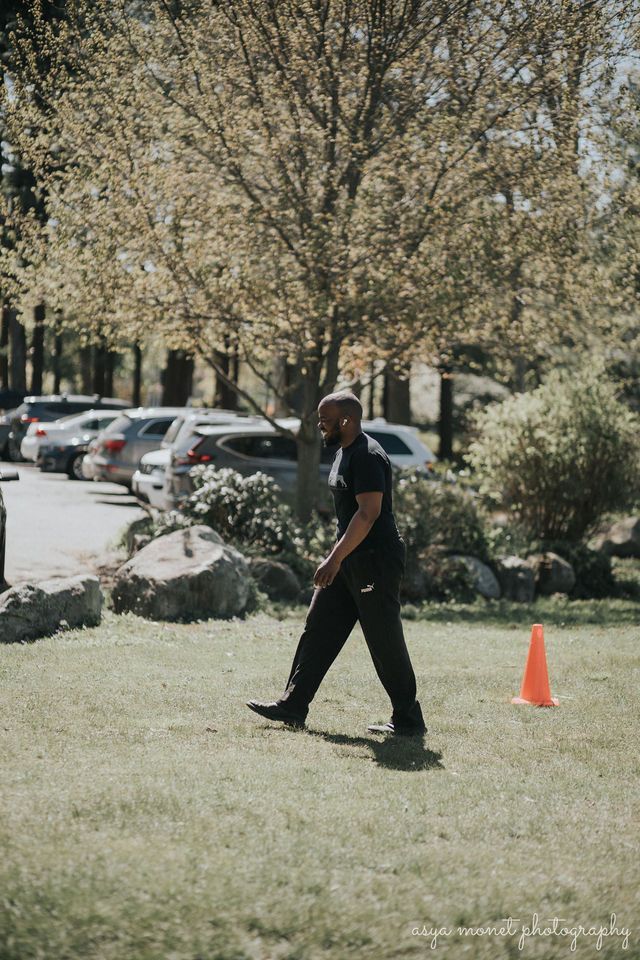 Person walking on a grassy field near an orange cone and parked cars under blooming trees.