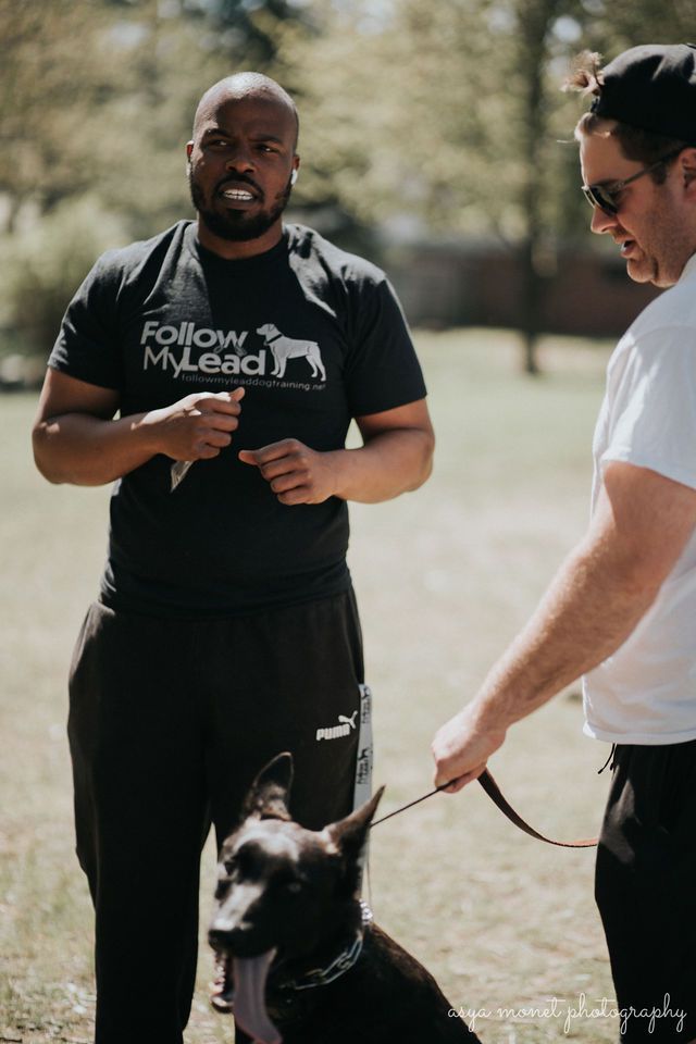 Man in black shirt holding leash with black dog beside another person outdoors
