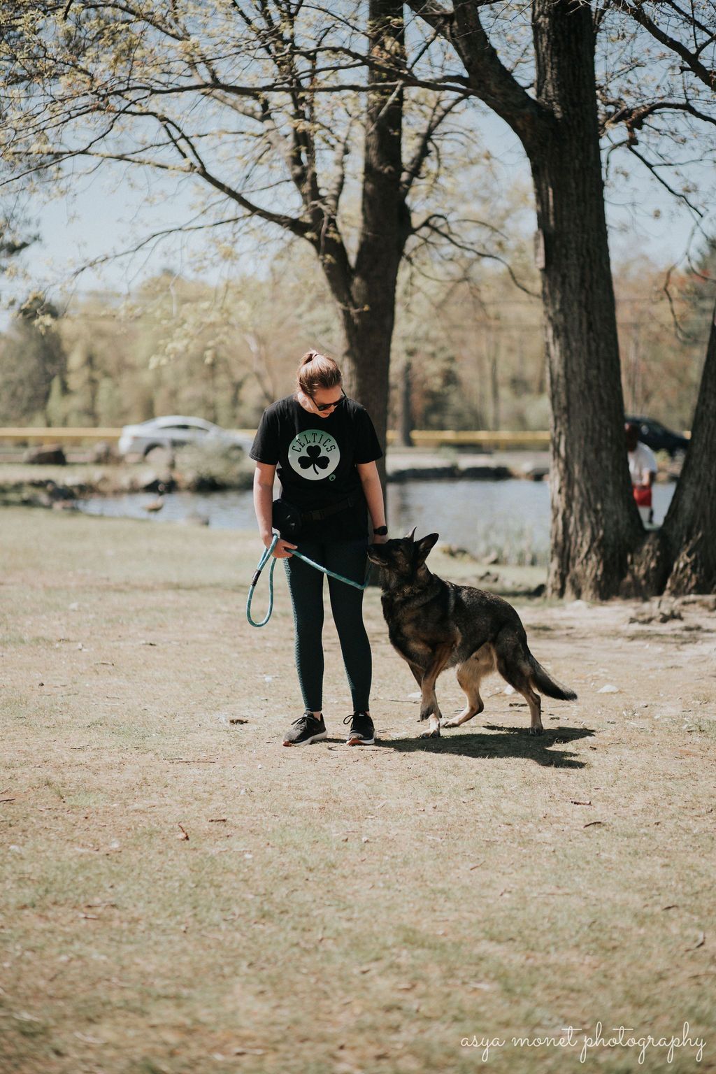 Person in black shirt stands with a German shepherd on a leash in a park under trees.