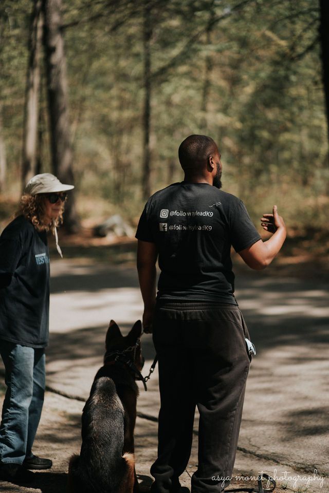 Trainer with a black dog and another person on a wooded path, both facing left.